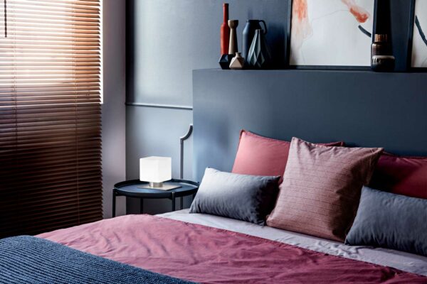 close-up-of-a-double-bed-decorated-with-pillows-in-a-hotel-room-interior-with-grey-wall-lamp-vases-and-window-blinds